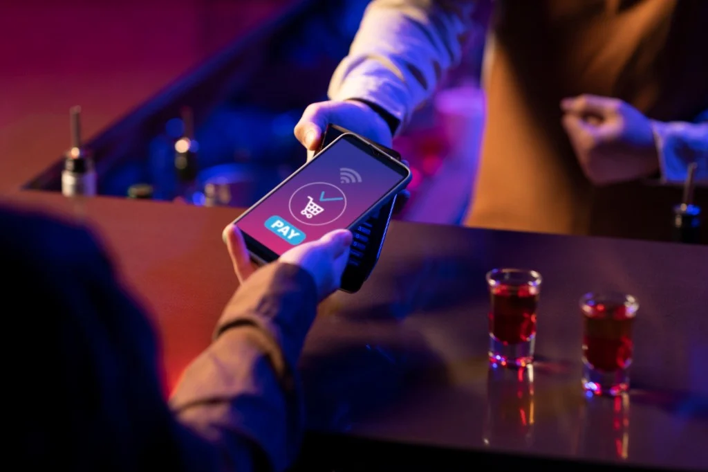 Customer paying with a smartphone on a POS terminal at a bar counter; neon purple and blue lighting; contactless payment screen with pay confirmation